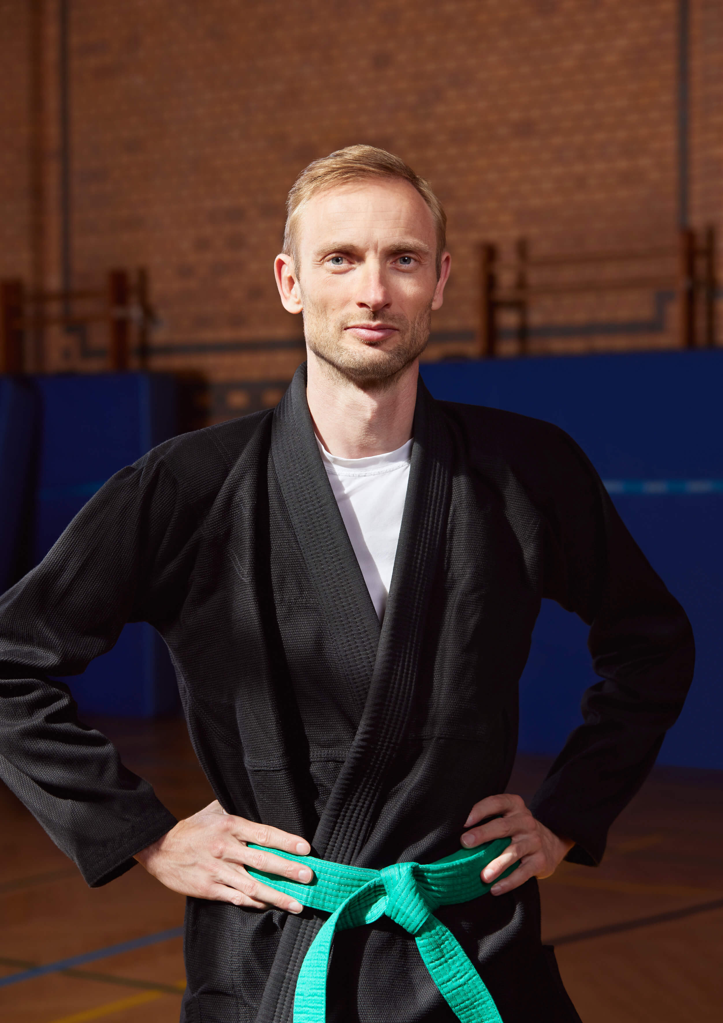 Eine Person mit einem Ju Jutsu Anzug, grünen Gürtel und blonden Haaren steht in einem Trainingsraum vor blauen Fußmatten und schaut in die Kamera.